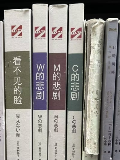 图片展示竖直排列的书脊特写，左侧四本为TLA出版社书籍：绿色封面《看不见的脸》（日文“見えない顔”）、紫色《W的悲剧》（“Wの悲劇”）、棕色《M的悲剧》（“Mの悲劇”）、深绿《C的悲剧》（“Cの悲劇”），均有红色TLA标志。右侧为其他书籍，含封面磨损的日文旧书及疑似村上春树作品《且听风吟》（书脊可见“Haruki”及“The Singing Wind”字样）。书籍整体有使用痕迹，书脊陈旧，呈现复古收藏感。