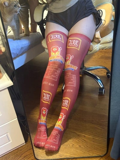 图片展示了一位坐在椅子上的人，穿着印有零食包装图案的红色过膝袜。袜子上有醒目的品牌标识、狮子吉祥物图案以及营养成分表等文字信息。背景中可以看到一只猫站在床上，好奇地望向镜头方向。整体场景是在一个居家环境中，有床、椅子和一些家具陈设。