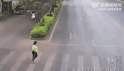 这是一段拍摄于广西柳州的动态画面，画面中一名身穿荧光绿上衣、黑色裤子的女子正在城市道路的斑马线上奔跑，画面左侧的非机动车道上有一辆电动车正在行驶，旁边是绿化带与停放的机动车，呈现出城市日常的道路场景。