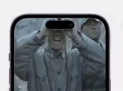 这是一张趣味二创梗图，画面主体是带灵动岛挖孔设计的iPhone正面，屏幕显示抗战剧《亮剑》中李云龙举望远镜观测的经典画面，iPhone顶部的黑色灵动岛挖孔恰好完美对齐望远镜两个镜筒之间的空隙，视觉上形成了李云龙正透过手机屏幕挖孔向外看的错位效果，创意十足很有幽默感。