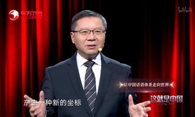 这是东方卫视政论节目《这就是中国》的视频截图，学者张维为身着深色西装、佩戴眼镜，正展开双手进行演讲，背景是红色幕布。画面配有演讲字幕“产生一种新的坐标”，还有主题提示文字“让中国话语体系走向世界”，整体场景严肃正式，是围绕中国国际传播话题的政论演讲内容，图中无谐音梗、双关或暗示内容。