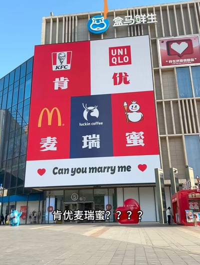 这是一张极具创意的商场外墙广告牌照片，通过肯德基、优衣库、麦当劳、瑞幸咖啡、蜜雪冰城五家品牌的logo和汉字，拼成了“肯优麦瑞蜜”，与英文“Can you marry me”谐音，用于表达求婚的浪漫场景。同时，下方有网友配文“肯优麦瑞蜜？？？”，表现看图后的搞笑、惊讶之情。整体画面体现了网络流行的品牌谐音梗文化，土味又可爱，适合用在求婚、表白等轻松娱乐话题。