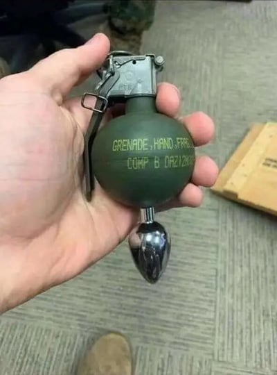 一只手握着一枚绿色的手榴弹，弹体上印有'GRENADE, HAND, FRAG'和'COHP B DRE1200'字样。手榴弹顶部有金属保险栓和拉环，整体呈现军用装备的特征。背景是室内环境，能看到地毯地板和模糊的纸箱。