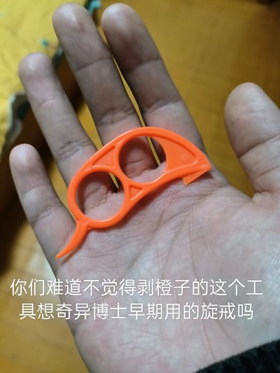 这是一张竖版实拍照片，画面主体是一只手掌托着一个橙色的双指环式剥橙器，该工具设计为双指套入的结构，一端带有尖状凸起用于插入橙皮，另一端有开口结构可辅助撬动橙皮，是常见的剥橙子小工具。图片下方配有白色文字，将这个日常工具与漫威超级英雄奇异博士早期使用的悬戒做趣味对比，调侃二者外形相似，为普通的生活小物品赋予了趣味的影视联想。
