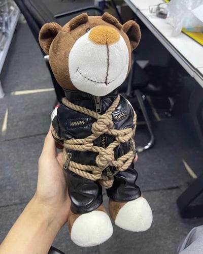 一只棕色的小熊玩偶，穿着黑色皮衣，被粗麻绳以复杂方式捆绑着，脸上却带着微笑。玩偶被一只人手握着，背景是办公环境，有桌椅等办公用品。这种反差萌的组合创造出一种幽默效果 - 凶狠的捆绑造型与可爱的微笑表情形成鲜明对比。