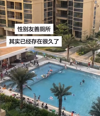 这是一张俯拍视角的高层住宅小区露天游泳池照片，背景是米黄色的高层住宅楼，泳池周边种植了棕榈树绿化，不少人在泳池边坐着休憩等候，泳池内有不少民众正在游泳玩乐，泳池被一道金属栅栏分隔成了两个独立区域；画面上方添加了两行白底黑字的配文，内容为“性别友善厕所 其实已经存在很久了”，玩梗调侃将这个被分隔开的游泳池戏称为早就存在的性别友善厕所，制造出搞笑效果。