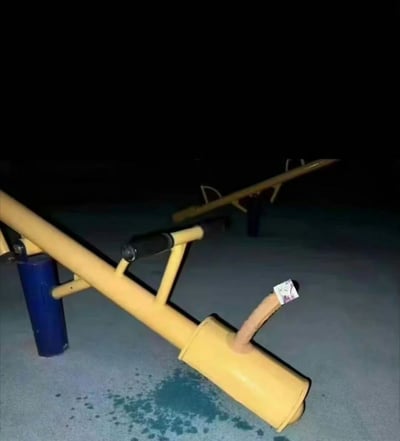 夜晚的游乐场场景，画面中央是一个黄色的秋千，秋千的座椅上贴着一张小纸片。秋千下方的地面上有一滩明显的蓝色液体污渍，背景较暗，只能隐约看到其他游乐设施的轮廓。整个场景在夜间灯光下呈现出一种神秘的氛围，蓝色污渍与黄色秋千形成鲜明对比，引人猜测污渍的来源。