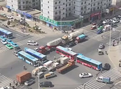 这是一张高空俯拍的城市十字路口实景照片，该路口各个方向驶入的多辆公交车、油罐车、货运卡车、出租车、私家车等机动车因抢行互不相让，全部挤在路口中央完全堵死，形成了近似环形的拥堵圈，路口周边分布有临街商铺和高层居民楼，可见少量行人和非机动车在路口周边活动，整体交通秩序完全瘫痪。