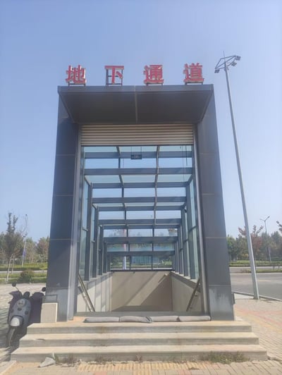 这是一张晴朗白天拍摄的城市户外地下通道入口照片。入口顶部设有醒目的红色立体字标识“地下通道”，入口区域是带有玻璃顶棚的下行通道，通道前有几级台阶，左侧停放着一辆灰色电动车。背景可见整洁的道路、绿化带和多臂路灯，整体呈现出城市公共设施的日常场景，环境开阔整洁。