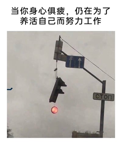 这是一张图文结合的职场类搞笑梗图，顶部配有文字“当你身心俱疲，仍在为了养活自己而努力工作”，下方是一个出现故障的交通信号灯：信号灯主体歪斜着下垂，仅红灯部分还悬挂着并保持亮起状态，整体形态如同一个疲惫不堪、即将支撑不住但仍在“坚持履职”的人。这张图用故障信号灯的状态，生动比喻了当代打工人在身心俱疲的情况下，为了生计不得不坚持工作的处境，精准戳中职场人的疲惫心态，引发共鸣。图中还有俄语“СТОП”（停止）标识和直行双车道指示牌，显示这是在俄语地区拍摄的场景。