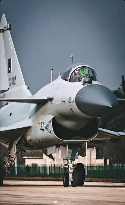 这张图片展示了一架巴基斯坦空军的歼-10CE战斗机停在机场跑道上的场景。战机机头正对镜头，驾驶舱内可见佩戴绿色飞行面罩的飞行员；尾翼印有巴基斯坦国旗的星月标志与编号“22-102”，机身的DSI进气道、前起落架等细节清晰呈现。背景是机场的围栏与低矮建筑，整体画面质感硬朗，凸显了战机的威严与专业性。