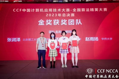 这是一张2023年CCF中国计算机应用技术大赛-全国算法精英大赛总决赛的金奖团队领奖现场照片。画面中共有四人站在舞台上，左侧是一位身着灰色POLO衫、深蓝色长裤的男性，推测为赛事嘉宾或获奖团队的指导老师；右侧三位是金奖团队“裙同”的成员，她们身着JK风格制服，佩戴口罩，手持红色获奖证书，站姿端正。背景是一块红色的大型显示屏，上面清晰标注了赛事名称、赛事年份、“金奖获奖团队”字样，还显示了部分团队成员的姓名与所属院校：北京理工大学的张润泽、华侨大学的赵雨扬，以及显示不全的团队名称“裙同”。屏幕右下角带有“CCF NCCA 中国计算机应用大会”的标识，整个场景正式庄重，记录了获奖团队收获荣誉的时刻。