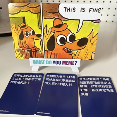 图片展示了"What Do You Meme?"派对游戏中的一张卡牌，卡牌上印有经典网络迷因"This Is Fine"的卡通狗形象。狗戴着帽子，身处着火的房间中，却微笑着说"THIS IS FINE"。卡牌下方有三张蓝色的中文 caption 卡片，内容分别是："当葬礼上的菜太好吃了以至于你都忘了是在吃谁的席了"、"当你因为长得不好看只好靠哗众取宠来吸引大家的注意力时"和"当他问你拿这个马桶刷是要刷墙还是刷马桶时，你意识到自己好像一直在用它洗澡来着"。
