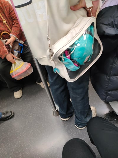 这是一张在地铁车厢内拍摄的日常通勤照片，画面中心的乘客背着一个米白色透明痛包，包内放置着蓝绿色的初音未来毛绒玩偶，拉链处还挂有同款形象的挂饰。画面左侧有拿着手机和塑料袋的乘客，右侧是穿着黑色羽绒服的乘客，整体呈现出普通的城市地铁通勤场景，没有双关或谐音梗内容。