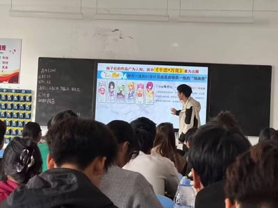 在一间高校教室内，一名学生站在讲台上，通过投影屏幕向台下众多同学介绍日本美少女游戏制作公司柚子社的代表作《千恋*万花》。屏幕上展示了游戏的六位角色立绘，以及游戏的特色“多重角色线路”相关介绍，提到游戏全线路通关需50-60小时，还出现网络热词“XP”指代个人喜好。教室黑板上记录着该班级的考勤信息，台下学生正在认真观看展示，整体是充满趣味的课堂展示场景。