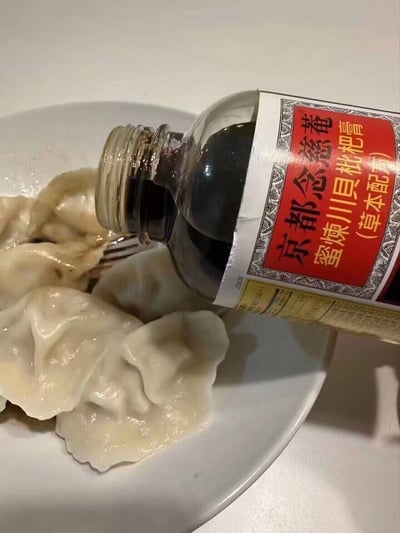 一张搞笑图片，展示了一瓶京都念慈菴蜜炼川贝枇杷膏被倒在一盘饺子上的场景。原本用于缓解咳嗽的传统草本糖浆被当作饺子蘸料使用，形成了有趣的错位搭配，产生幽默效果。