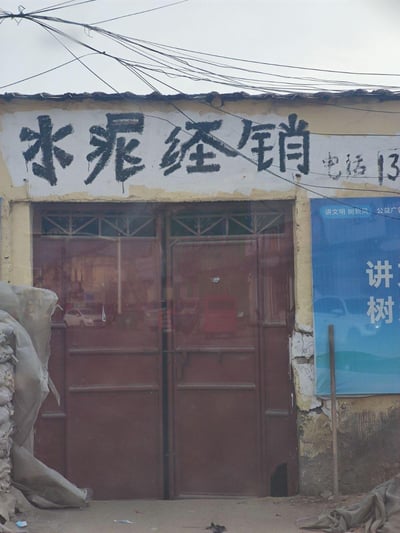 一张展示老式水泥经销店门面的照片。照片中可以看到一个略显陈旧的店铺，墙面有些斑驳，上方有手写的黑色大字“水泥经销”，旁边还有部分可见的电话号码“电话13”。店铺有两扇红色的玻璃门，门上有简单的花纹装饰。门的右侧贴着一张蓝色的公共宣传栏，上面有白色文字“讲文明 树新风”。店铺上方有许多杂乱的电线横跨，整体呈现出一种朴实、怀旧的氛围，反映了小型传统生意的风貌。