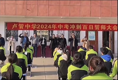 卢管中学2024年中考冲刺百日誓师大会现场，学生们穿着黄黑相间的校服整齐坐在前方，舞台上站着多位老师或工作人员，他们正举起右手进行宣誓。背景是学校的红砖墙面，上方悬挂着醒目的红色横幅，写有“卢管中学2024年中考冲刺百日誓师大会”字样，整体氛围庄重而充满激励感。