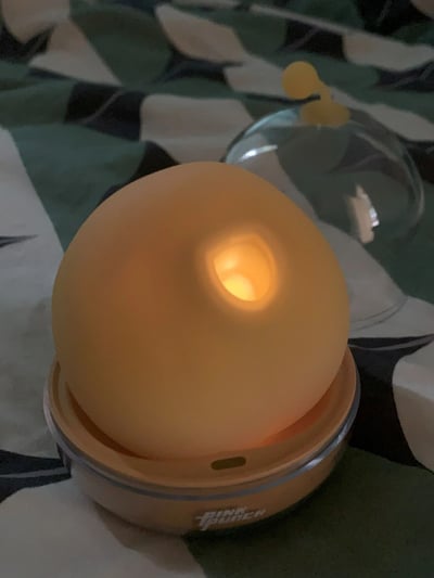 这是一张原始拍摄的静态竖图，主体是一个正在发光的球形暖光小夜灯。小夜灯的球体为磨砂质感的淡黄色，正面有一个凹陷区域，散发出柔和的暖黄色光线，营造出温馨的氛围。小夜灯的底座是浅棕色，带有金属质感的边缘，底座正面印有白色的“PINK PUNCH”品牌字样。背景是带有绿、白、黑几何图案的布料，在小夜灯的后方还放置着一个透明的半球形罩子，罩子顶部带有一个黄色小部件。
