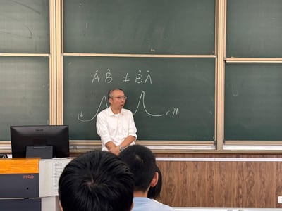 这是在一间大学教室的授课场景，一位戴眼镜的男老师站在绿色拼接黑板前，正在讲解量子力学知识。黑板上写着量子力学核心的不对易关系公式$ÂÂAÂÂB ≠ ÂÂBÂÂA$，还手绘了两个类似光谱的曲线，标注了数字“b”和“91”。前景能看到几位听课学生的后脑勺，讲台左侧摆放着一台飞利浦（PHILIPS）显示器，讲台带有“FOCUS”标识，整体氛围是严肃专注的大学专业课课堂，没有双关、谐音梗或暗示内容。