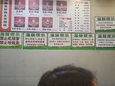 这是一张线下火锅店的店内实拍横图，画面上方是锦咖美蛙鱼头火锅店的价目菜单，列出了火锅鸡、凤爪鸡锅、排骨鸡锅等不同规格的锅品，以及单加菜品、凉菜、主食的对应售价。菜单下方并排张贴了多张绿白配色的“温馨提示”告示，内容一反常规提示的正式风格，全部是诙谐玩梗的免责声明，包含禁止顾客吃绿萝咬瓶盖、不提供拜把子道具、禁止酒后在店内随意约架、自带酒水需要家属确认、痔疮患者不建议点特辣、店内不存在冰红茶类饮品等荒诞又搞笑的内容，充满网感，画面底部还露出了拍摄者的部分头顶。