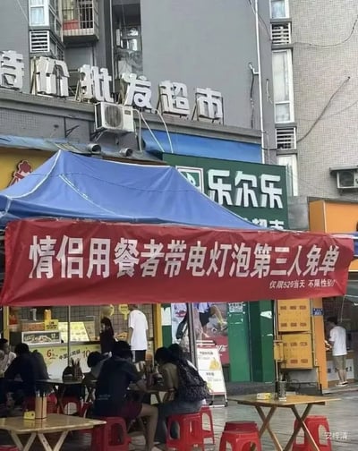 这张图片拍摄的是一家餐饮摊位，在其遮阳篷下悬挂着一幅横幅，内容为“情侣用餐者带电灯泡泡第三人免费”。这里的“电灯泡”原本指的是两人约会时出现的第三者（通常有点尴尬），而“泡”又和“泡面”等饮食场景相关，有明显的文字双关和谐音梗。图片现场有不少人在用餐，营造出一种轻松搞笑的气氛。横幅下方补充说明“仅限520当天 不限生和”，利用520（谐音“我爱你”）营造节日氛围。整体非常适合社交媒体传播，调侃情侣和单身人士的社交尴尬，很有互联网恶搞气质。

本图包含典型的双关和谐音梗，“电灯泡”既指多余的第三人，也暗含饮食中的“泡”。