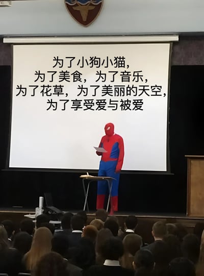 图片中，一名身着蜘蛛侠红蓝经典 costume 的人站在讲台上，手持纸张正在进行演讲。讲台后方的投影屏幕上显示着中文文字：“为了小狗小猫，为了美食，为了音乐，为了花草，为了美丽的天空，为了享受爱与被爱”，内容充满对生活美好事物的向往。台下坐着众多听众，背景上方有一个带有动物图案的纹章装饰，整体场景像是在学校礼堂或类似的公共演讲场所。