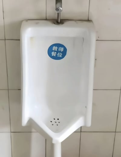 这是一张实拍的公共男卫生间场景图，画面主体是安装在米白色瓷砖墙面上的白色立式小便池，小便池正面显眼位置贴有一个蓝色圆形标识，标识上印有白色的“教师餐位”四个汉字，原本属于食堂的座位标识被张贴到小便池上，形成了强烈的反差感，极具搞笑讽刺效果。