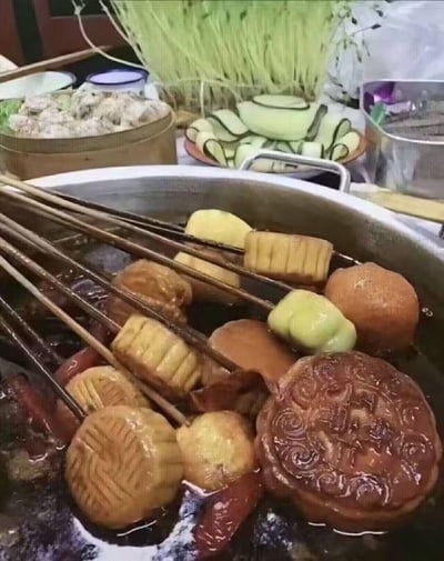 这张图片展示了一锅正在煮的串串火锅，其中除了常见的面点、丸子类串串外，还混入了广式月饼和造型类似月饼的面点串串，将传统中秋食品月饼以完全颠覆常规的方式——涮火锅来呈现，属于典型的创意黑暗料理，画面充满了滑稽感，打破了人们对月饼食用场景的固有认知，用来调侃中秋月饼的另类吃法，制造出新奇搞笑的效果。