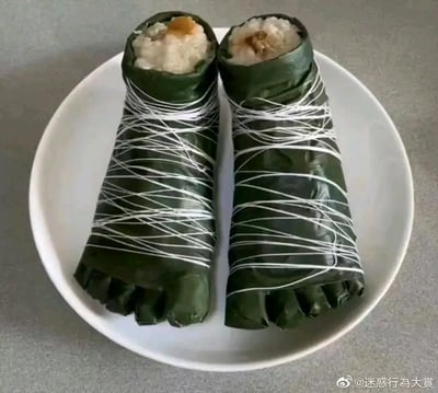 这张静态单图展示了两只被制作成人脚形状的创意粽子，放置在一个白色圆形盘子中。粽子以粽叶为“外皮”，底部被塑分出脚趾的形态，外层用白色棉线交叉缠绕固定，顶部的粽叶被打开，露出混合了馅料的糯米，仿佛是脚的前端被掰开露出内部。这种打破传统的趣味造型，让原本普通的粽子变得极具视觉冲击力，制造出搞笑、新奇的效果，没有双关或谐音梗，属于创意美食的范畴。