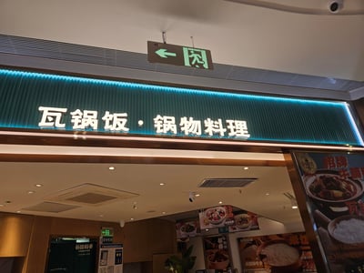 这是一张谷田稻香连锁中式快餐的门店实拍图，门店招牌为「瓦锅饭·锅物料理」，搭配蓝色背光设计十分醒目。店内天花板可见安全出口指示牌与监控设备，同时悬挂着招牌牛腩锅、番茄鱼等菜品的宣传海报，还有9.9元米饭畅吃卡的活动信息，门口处设有蜀山区食品安全监管信息公示栏，整体呈现出商场内连锁快餐门店的日常场景。