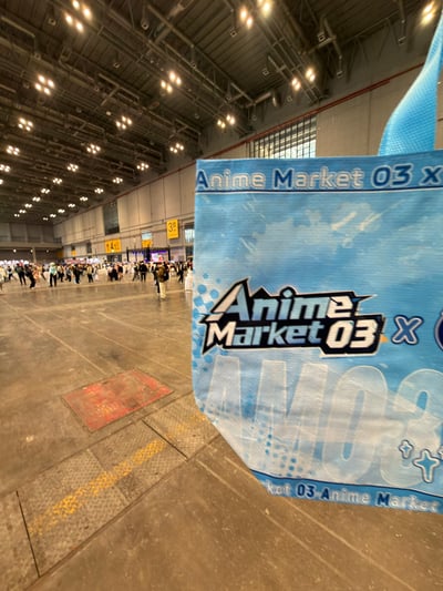 这是一张实拍照片，拍摄于大型室内会展场馆内部。前景主体是一个浅蓝色的无纺布袋，袋身印有醒目的「Anime Market 03」字样，是该届动漫展会的周边物料。背景是宽敞开阔的展馆空间，地面为水泥质地，远处有大量参展人员走动，展馆墙面上悬挂着标有3、4号馆的黄色指示标识，场馆顶部排布着密集的照明顶灯，整体呈现出第三届Anime Market动漫展会在上海举办的现场场景。