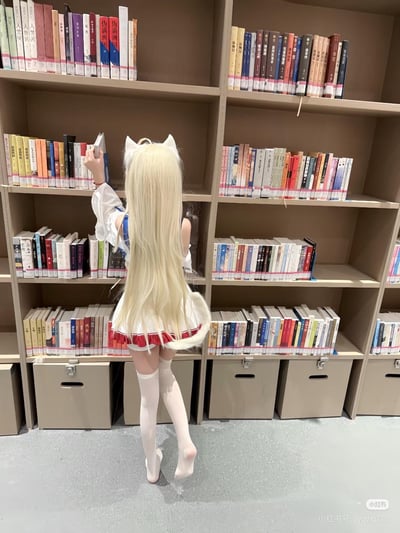 一位身着猫耳cos服装、留着蓬松金色长发的女生背对着镜头，站在摆满各类中文书籍的木质书架前，正抬手从第二层书架上取书。她的服装为白红配色，搭配白色过膝长袜，还带有猫尾装饰，整体场景将二次元的可爱风格和图书馆的文艺氛围结合在一起，显得温馨又治愈。