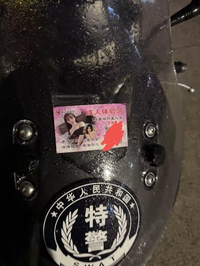 这张实拍图呈现了极具反差感的场景：一张标注「18+禁止未成年」的成人体验馆小广告贴纸，被贴在带有「中华人民共和国 特警」标识的黑色装备表面，装备上还有水渍显得潮湿。广告贴有女性模特图片，宣传「1:1高端仿真人偶」，并标注「干净卫生 配套齐全 合法经营 安全放心」，附带的联系方式/二维码被红色涂抹遮盖，整体画面充满荒诞感。