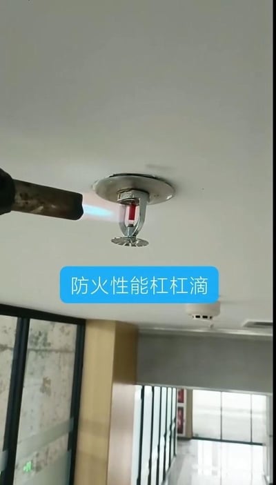 图片展示了一个天花板上的防火喷淋头，左侧有一个金属管（疑似喷火器）正对着喷淋头喷射火焰，下方有蓝色文字标注“防火性能杠杠滴”。场景可能是在办公室或商业建筑内进行的防火性能测试，通过实际火焰测试喷淋头的耐高温或防火效果，整体画面清晰，突出消防安全设备的可靠性。