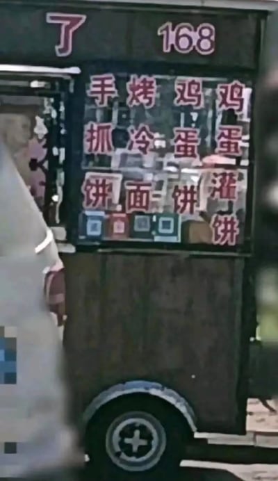 这是一张街头流动小吃餐车的实拍图，餐车窗口以竖排红色手写字体展示了四款主营小吃：手抓饼、烤冷面、鸡蛋饼、鸡蛋灌饼，窗口顶部还有单独的“了”字和数字“168”，窗口下方贴有四个不同平台的收款二维码。竖排的排版方式让菜单乍看容易产生有趣的误读组合，充满了市井烟火气，这类餐车通常在居民区、学校周边提供平价便捷的早餐或简餐。
