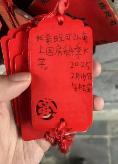 一只手拿着几张红色的许愿牌，其中最前面的一张写着：'我希望旺可以考上国防科技大学。2025年2月15日 钟旺'。这些许愿牌有着传统的中国设计，底部有一个镂空的'福'字图案。背景中可以看到更多类似的红色许愿牌，暗示这是在某个寺庙或许愿场所拍摄的照片。