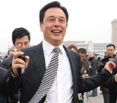 这是一张经过换脸P图改编的照片，将特斯拉CEO埃隆·马斯克的面部合成到一位身着西装领带的中国男士身体上。该男士处于一个类似公开活动的场景中，周围环绕着不少手持拍摄设备的人员，背景可见具有标志性的城市建筑。由于面部与身体的违和拼接，整体画面呈现出滑稽搞笑的效果，属于网友创作的趣味二次创作内容。