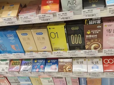 这是一张超市计生用品区货架的实拍照片，货架上整齐陈列着杜蕾斯、杰士邦、冈本等多个知名品牌的不同款式安全套，每个商品下方都对应有标注价格的价签。其中一款黄色包装的安全套设计特殊，印有大号黑色数字「01」以及「白名单凭证1.0」的文字，其外观与手游《明日方舟》中的白名单凭证道具UI高度相似，结合发布者配的标题「明日方舟还在追我」形成玩梗效果，调侃在现实生活中也能偶遇游戏相关的元素，仿佛游戏一直跟着自己。