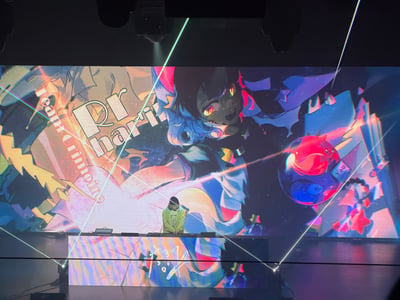 这是一张横向拍摄的室内电子音乐现场照片。画面中央偏下位置，一名穿荧光绿色连帽衫、戴帽子的DJ正低头操作台前的设备。DJ身后是一块几乎占据整个背景的巨型LED屏，屏上播放着色彩浓烈的动漫风视觉图：一名蓝红相间发色、橙黄发光眼睛的角色向前伸手，周围有破碎几何图形、星星、卡牌和红蓝透明球体等元素。屏幕左侧可见竖排的“team Grimoire”字样，中部偏左有大号标题文字，结合曲师信息可判断为《Rrhar'il》，部分文字被激光束遮挡。舞台上方可见射灯和吊装设备，多束绿、白、紫等颜色的激光从画面中穿过，DJ台正面也有炫彩条纹和白色手写风格图案，整体氛围迷幻、高能，具有典型的音游曲或硬核电子音乐演出特征。