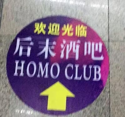 这是一张拍摄于地面的圆形商家指引标识，标识底色为紫粉渐变，从上到下依次是黄色的「欢迎光临」字样、白色的「后末酒吧」字样、白色的英文「HOMO CLUB」，最底部配有一个黄色的向上箭头，用于指引顾客前往酒吧的方向。值得注意的是，英文名称中的「HOMO」在英文俚语中常被用来指代同性恋者，与中文名称「后末酒吧」形成了谐音巧合，带有一定的趣味梗，容易引发观者的联想。