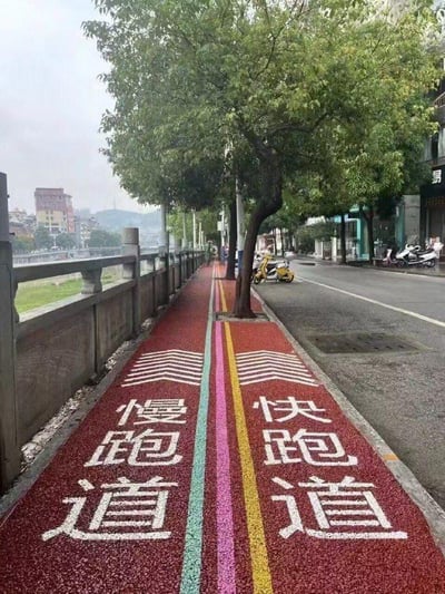 这是一张城市滨江健身步道的实拍图，步道采用红色透水路面，中间以绿、粉、黄三色线条划分区域，左侧清晰标注“慢跑道”，右侧标注“快跑道”，区域上方还有向前的箭头标识，用于区分不同跑步节奏的健身人群。步道一侧是江边护栏，可眺望远处的江水与城市建筑，另一侧是城市街道，路边停放着电动车，行道树间隔分布在步道上，整体是为满足不同健身需求而设计的人性化城市便民设施。