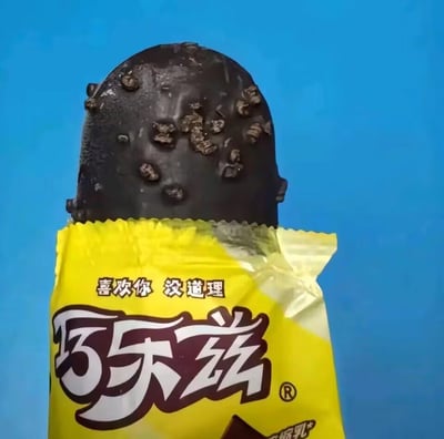 这是一张巧乐兹品牌冰淇淋的实拍图，冰淇淋外层裹着带有碎粒的深棕色巧克力脆皮，部分亮黄色的品牌包装被剥开，包装上印着品牌名“巧乐兹”和宣传语“喜欢你 没道理”，背景是纯净的蓝色，清晰展示了冰淇淋的外观，看起来口感浓郁，充满夏日解暑的吸引力。图中没有双关、谐音梗或暗示内容。