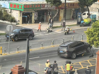 这是一张城市街道的实拍照片，画面中展示了临街的多家店铺，包括绿色招牌的“敏迪化工”、红色招牌的“永安广告”以及“天蓬门业”等，店铺门口有行人走动。街道上有黑色SUV、摩托车和电动三轮车等交通工具，一名身穿橙色工作服的人员正在过马路，另有几位路人驻足观望。右侧一辆黑色SUV前轮骑在道路中央的隔离护栏上，疑似发生了小型交通意外，周围有穿着反光背心的人员在场，整体场景充满生活气息与即时性。