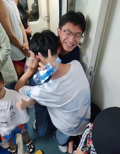 这张图片拍摄于火车车厢的过道处，一位年轻人靠在门边半跪休息，手里拿着一瓶水，身边有穿着夏装的小孩和穿花衬衣的成年人，大家在炎热的环境下感到明显的疲惫。由于没有座位，一家人只能挤在狭小的空间中，展现出真实的中国暑期大众交通、家庭出行的压力画面。整体氛围真实、无奈、贴近日常。

本图没有明显双关、谐音梗或暗示内容。