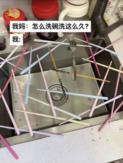 整张图拍摄的是厨房水槽的实景，水槽内横七竖八散落了多根颜色各异的塑料筷子，还有一把金属叉子立在筷子中间，整体布局完全复刻童年经典玩具「挑竹签」的游戏开局状态。图片上方配有两行文字对话，第一行是「我妈：怎么洗碗洗这么久？」，第二行是「我:」后无文字，用画面生动展现出「我」本该洗碗却在水槽玩起了挑筷子的游戏，因此耽误了时间的摸鱼状态，笑点自然，容易引发网友对做家务时偷偷摸鱼的共鸣。