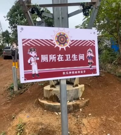 这是一张户外实拍照片，拍摄于攸乐神秘雨林徒步路线的户外区域，地面为红土路面，背景可见植被、金属支撑架，远处还有配备户外装备的人员与车辆。画面核心是固定在金属杆上的指示牌，牌面以红白色为主色调，顶部有民族特色的太阳形纹饰，左右两侧分别绘制身着少数民族服饰的卡通男女形象，牌面中央印有白色大字“厕所在卫生间”，底部标注有“攸乐神秘雨林徒步”字样，该标语属于逻辑重复的废话式表述，颇具荒诞搞笑的效果。