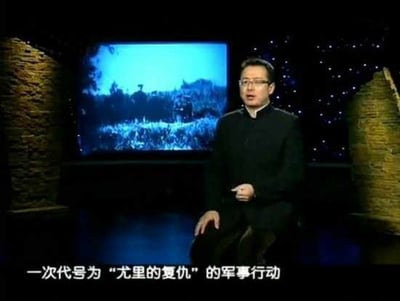 这是一张军事纪实类节目的视频截图，画面中一位戴眼镜、身着黑色中式立领服装的主持人端坐于演播室中，背景屏幕播放着带有士兵形象的黑白历史军事影像，画面底部配有字幕：“一次代号为‘尤里的复仇’的军事行动”，整体场景是主持人在为观众讲解相关军事行动的内容。