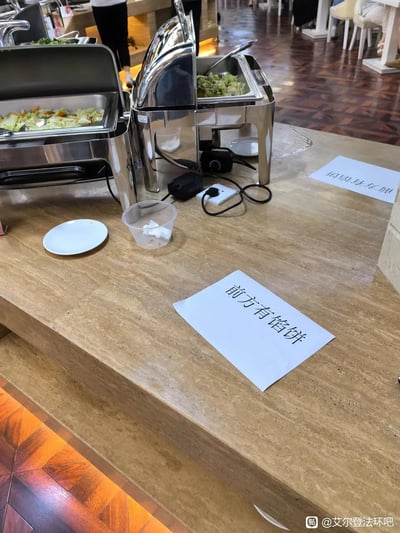 这是一张在自助餐厅拍摄的实拍图，餐台上摆放着装有蔬菜餐品的保温餐炉，台面上散落着空白盘、装有纸巾的透明小塑料盒，还有两张写着「前方有馅饼」的白纸。背景能看到餐厅的拼花地板和其他模糊的顾客身影。这是玩了游戏《艾尔登法环》中「前方有陷阱」的谐音梗，用「馅饼」替代「陷阱」，在餐厅场景里制造出趣味整活的效果，调侃感拉满。