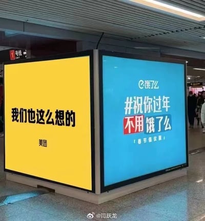 这张图片拍摄于地铁站内，画面主体是相邻的两个方形广告位。左侧是美团的黄色背景广告，上面用黑色字体写着“我们也这么想的”；右侧是饿了么的蓝色背景广告，内容为“#祝你过年不用饿了么 「春节倡议版」”。两个品牌的广告形成了趣味联动，饿了么提出春节不用点外卖的倡议，美团则表示认同，传递出鼓励大众在春节期间回归家庭、享用家常餐食的暖心氛围，图中没有双关、谐音梗等内容。背景中可以看到地铁站的1号线标识和行人，显示出这是一个人流量较大的公共交通场景。